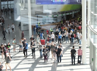 A large group of people gathered in a spacious, well-lit indoor area with glass walls. Many individuals are moving around, some in conversation and others holding bags. There is a large, visible queue in the background, organized by barriers, and several people are wearing gaming-related apparel. Signs with text hang from the ceiling.