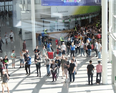 A large group of people gathered in a spacious, well-lit indoor area with glass walls. Many individuals are moving around, some in conversation and others holding bags. There is a large, visible queue in the background, organized by barriers, and several people are wearing gaming-related apparel. Signs with text hang from the ceiling.