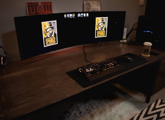 A gaming setup featuring a large curved monitor displaying PUBG logos on the screens. A keyboard and mouse are positioned on a large desk pad. A pair of headphones rests on the desk alongside a mobile phone. A drink in a Starbucks cup is placed to the right, while a desktop tower is seen in the background on the right side.