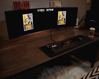 A gaming setup featuring a large curved monitor displaying PUBG logos on the screens. A keyboard and mouse are positioned on a large desk pad. A pair of headphones rests on the desk alongside a mobile phone. A drink in a Starbucks cup is placed to the right, while a desktop tower is seen in the background on the right side.