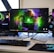A gaming setup featuring a central monitor with a vibrant display showing 'The Spirit of Gaming'. On either side, there are gaming PCs with illuminated components and transparent cases. Headsets hang from the side of the monitor. The desk also has a black keyboard and mouse, and a branded mousepad.