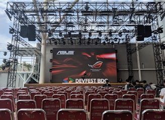 A stage setup with a large screen displaying logos for ASUS and Republic of Gamers. Rows of red chairs are arranged in front of the stage. The event is titled 'DEVFEST BDC', suggesting a tech or gaming conference. The venue appears to be indoors with a visible ceiling truss structure.