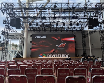 A stage setup with a large screen displaying logos for ASUS and Republic of Gamers. Rows of red chairs are arranged in front of the stage. The event is titled 'DEVFEST BDC', suggesting a tech or gaming conference. The venue appears to be indoors with a visible ceiling truss structure.