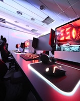 A row of high-end gaming computers on sleek desks, featuring RGB lighting and gaming accessories. The computers are housed in transparent cases with visible components, emitting a red glow. Comfortable gaming chairs with ergonomic design are positioned next to the desks, and the room's ceiling is modern with recessed lighting.