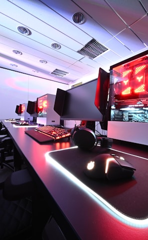 A row of high-end gaming computers on sleek desks, featuring RGB lighting and gaming accessories. The computers are housed in transparent cases with visible components, emitting a red glow. Comfortable gaming chairs with ergonomic design are positioned next to the desks, and the room's ceiling is modern with recessed lighting.
