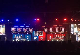 An esports event with a large screen displaying two teams, Team Kaliber and Rise Nation, competing against each other. The screen shows profiles of team members and scores, with Team Kaliber leading 2-0. The venue is dimly lit with blue and red lighting, and an audience can be seen watching.