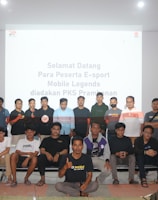 A group of people is posing for a photograph in a room with a white screen in the background that has text welcoming e-sport participants for a Mobile Legends event organized by PKS Prambanan. The group consists of 20 individuals standing and sitting, with some wearing casual attire and others wearing matching shirts that include logos.