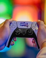 Two hands are holding a gaming controller labeled 'Pro Gamer'. The background is blurred with colorful lights creating a vibrant atmosphere.