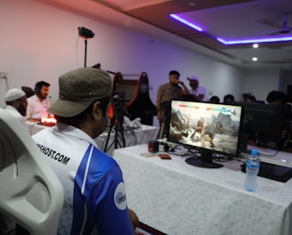 A group of people is gathered in a room watching and participating in a video game competition. Players and spectators are focused on the monitors displaying a fighting game. The setting includes tables with equipment and chairs arranged around the venue, with a noticeable intensity in the participants’ engagement.