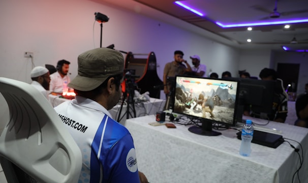 A group of people is gathered in a room watching and participating in a video game competition. Players and spectators are focused on the monitors displaying a fighting game. The setting includes tables with equipment and chairs arranged around the venue, with a noticeable intensity in the participants’ engagement.